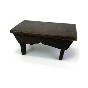 May include: A small, antique wooden bench with a dark brown finish. The bench has a rectangular top and four short legs. The wood shows signs of age and wear, adding to its vintage charm. The bench is approximately 12 inches long.