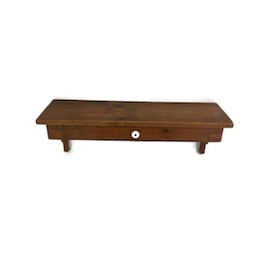 May include: A small, rectangular wooden shelf with a single drawer. The shelf is stained a warm brown color and has a white knob on the drawer. The shelf is supported by four short legs. A rustic, vintage-style piece.