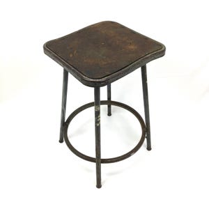 May include: An industrial-style metal stool with a square seat and four legs. The seat is a dark, weathered brown, and the legs and circular footrest are a matching dark gray. The stool has a vintage, rustic appearance.
