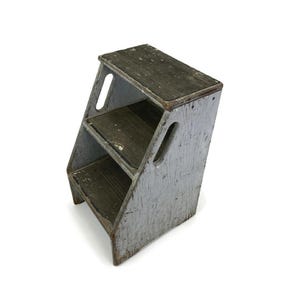 May include: A two-step wooden step stool with a distressed light gray painted finish. The stool features a rectangular top step and a lower step, both with dark, textured surfaces. Each side panel includes an elongated oval cutout handle.
