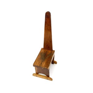 May include: A tall, narrow wooden chair with a single vertical backrest and a rectangular seat. The wood features a medium brown finish with a dark, hand-painted botanical illustration of wheat stalks on the seat surface and a light wood base.