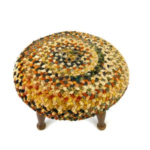 May include: A round, multi-colored footstool with a textured, woven surface. The color palette includes shades of orange, yellow, green, and black. The stool is supported by three short, dark wooden legs, creating a unique and decorative piece of furniture.