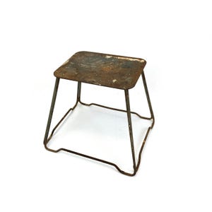 May include: A small, square metal stool with a weathered, rusted surface on the top platform. The stool features four thin, angled metal legs connected by a rectangular wire frame base, showing signs of age, oxidation, and industrial wear.