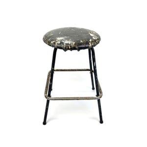 May include: A vintage, round-top stool with a distressed, black seat and a black metal frame. The seat shows signs of wear with white paint splatters. The stool has four legs and a footrest, perfect for a kitchen or bar setting.