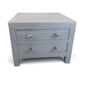 May include: A small gray wooden dresser with two drawers and glass knobs. The dresser has a simple design and is perfect for a small space.