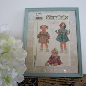 May include: A framed Simplicity sewing pattern for children's dresses. The pattern shows illustrations of three dress designs. The frame is a light blue colour, and the background is white.