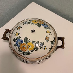 May include: Round, lidded casserole dish with a floral design. The lid and body are white with a silver rim. The floral pattern includes blue, yellow, and orange flowers with green leaves. The handles are brown.