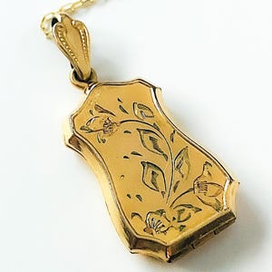 Op de afbeelding: Een goudkleurige rechthoekige hanger met bloemengravures, hangend aan een delicate gouden ketting. De hanger heeft een scharnierend ontwerp en ingewikkelde details, die een vintage esthetiek laten zien.