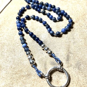 May include: A necklace featuring dark blue beads and silver-colored beads, connected by a blue cord. The necklace has a large silver-colored ring at the bottom. The beads are small and round, and the necklace is displayed on a light brown surface.