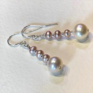May include: A pair of drop earrings featuring silver hooks, each dangling three small mauve-colored pearls followed by one larger white pearl. The pearls are separated by tiny silver beads, and the entire structure is connected by silver wire.