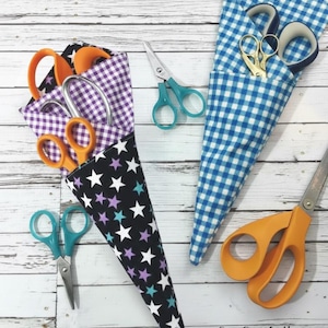May include: Scissors in various sizes and colors, including orange, teal, and gold, are displayed. They are stored in fabric cone-shaped holders with star and gingham patterns. The image is set on a white wooden surface.