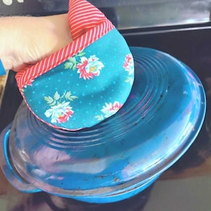 May include: A teal and red floral print oven mitt rests on a blue pot. The mitt features a teal fabric with white polka dots and pink and white floral designs. The top of the mitt is red and white striped.