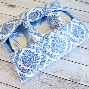 May include: An insulated lunch bag in a blue and white damask pattern. The bag has a handle and a light blue interior. The bag is open, revealing a silver insulated compartment.