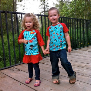 May include: Two children wearing colorful shirts with cartoon characters. The girl is wearing a blue and red shirt with a Mickey Mouse design. The boy is wearing a blue and red shirt with a cartoon character design. They are standing in front of a black metal fence.