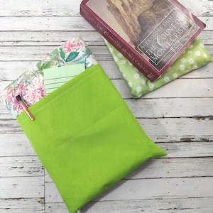 Puede incluir: Una funda de libro verde lima con un bolsillo floral y un bolígrafo. Una copia de "El Conde de Montecristo" descansa sobre una tela floral verde. La funda del libro está sobre una superficie de madera blanca.