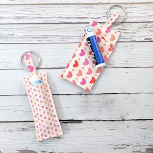 May include: Two fabric keychain holders with a lip balm inside. One holder has a pattern of pink and red hearts, and the other has a pattern of pink and orange crosses. Both have a silver key ring.