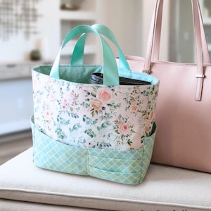 May include: A floral print tote bag with teal handles and pockets. The bag features a light pink and white floral pattern with green leaves. The bottom section and handles are teal. A pink leather tote bag is next to it.