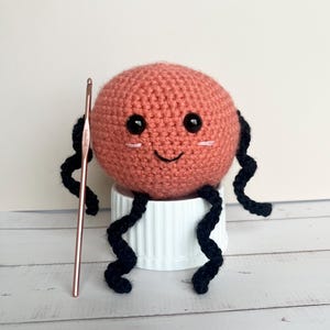 May include: A handmade, coral-colored crochet creature with black curly appendages and a smiling face. It has black button eyes and pink blush marks. A rose gold crochet hook is positioned next to the creature. The creature is sitting on a white, ribbed container.