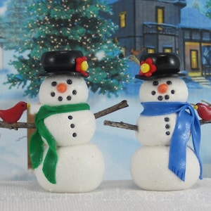 May include: Two miniature snowman figures, each with a black top hat, carrot nose, and stick arms. One wears a green scarf, the other a blue scarf. A red bird perches on a branch on each. Festive winter decoration.