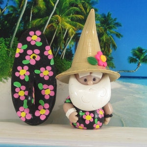 Peut inclure: Une figurine fantaisiste de gnome de plage avec une longue barbe blanche, portant un chapeau de paille et une chemise hawaïenne à fleurs roses et jaunes. À côté du gnome se trouve une planche de surf noire décorée de fleurs roses et jaunes.