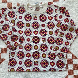 May include: White long-sleeved shirt with a red, yellow, and blue geometric pattern. The pattern features hearts, diamonds, and other shapes.