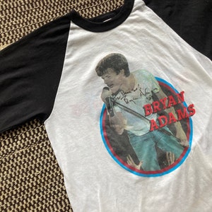 May include: A white and black baseball tee shirt with a red, white and blue circle graphic featuring a photo of Bryan Adams singing into a microphone. The text "Bryan Adams" is written in red letters on the circle. The shirt has a black signature on the graphic.