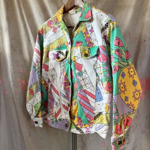 Puede incluir: Chaqueta estampada con botones y dos bolsillos en el pecho. La chaqueta presenta formas geométricas y diseños florales en blanco, rosa, verde, amarillo y morado. La chaqueta está colgada de una percha de madera.
