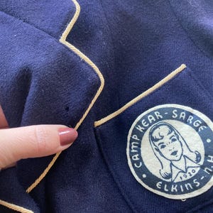 Może przedstawiać: Granatowa marynarka z złotą lam&oacute;wką i naszywką na lewej piersi. Naszywka jest biała z czarnym napisem "Camp Hear-Sarge Elkins-N.H. 1920". Naszywka zawiera r&oacute;wnież czarno-białą ilustrację dziewczyny z długimi włosami.