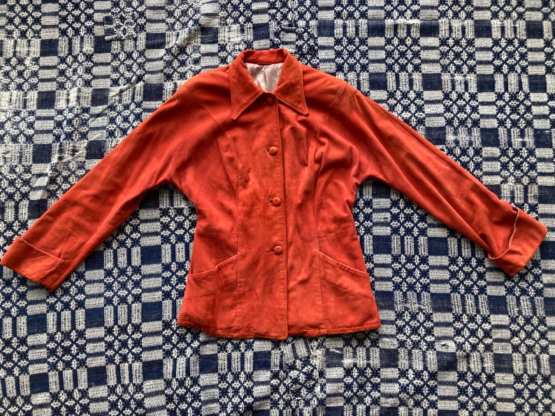 May include: A rust-colored suede jacket with a collared neckline, long sleeves, and button closure. The jacket has two front pockets and a fitted silhouette.