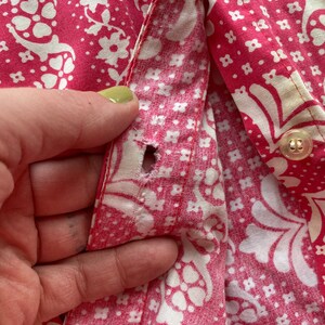 May include: A close-up of a pink and white patterned fabric with a button and a hole in the fabric. The fabric is likely from a shirt or blouse.