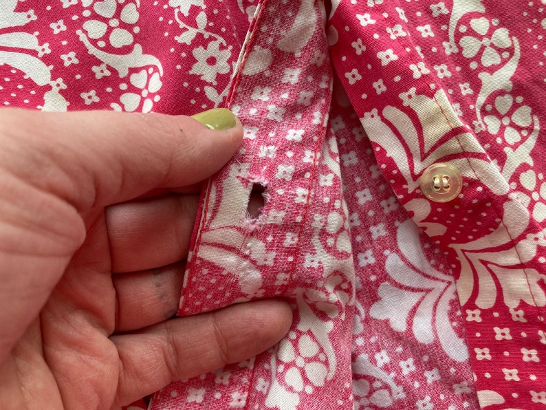 May include: A close-up of a pink and white patterned fabric with a button and a hole in the fabric. The fabric is likely from a shirt or blouse.