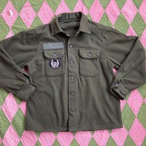 Peut inclure: Une chemise boutonnée à manches longues de couleur vert foncé avec un patch de crâne sur la poitrine gauche. La chemise a deux grandes poches sur le devant et un col boutonné.