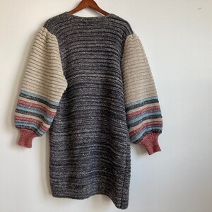 May include: A gray and beige striped knitted sweater with long sleeves and a v-neck. The sleeves have a multi-colored stripe pattern in pink, blue, and white.