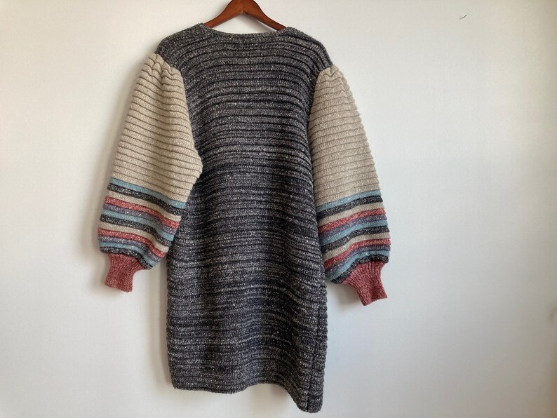 May include: A gray and beige striped knitted sweater with long sleeves and a v-neck. The sleeves have a multi-colored stripe pattern in pink, blue, and white.