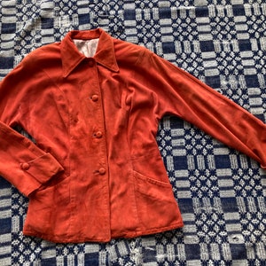 May include: A rust-colored suede jacket with a collared neckline, button closure, and long sleeves. The jacket has two pockets on the front and a slightly fitted silhouette.
