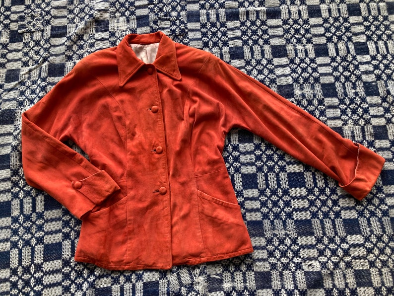 May include: A rust-colored suede jacket with a collared neckline, button closure, and long sleeves. The jacket has two pockets on the front and a slightly fitted silhouette.