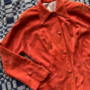 May include: A burnt orange suede jacket with a collared neckline and button closure. The jacket has two pockets and long sleeves with buttoned cuffs.