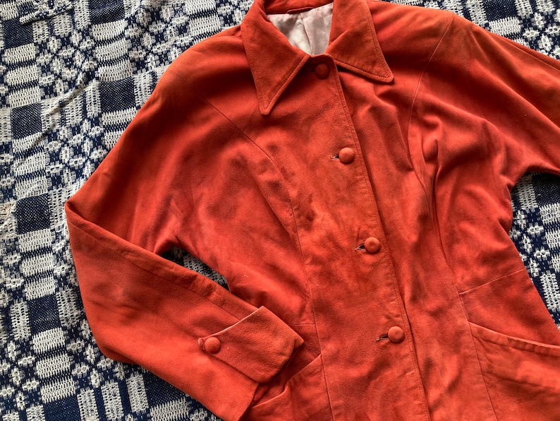 May include: A burnt orange suede jacket with a collared neckline and button closure. The jacket has two pockets and long sleeves with buttoned cuffs.