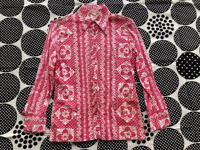 May include: A pink long-sleeve button-up shirt with a white floral pattern. The shirt has a pointed collar and a button-up front.