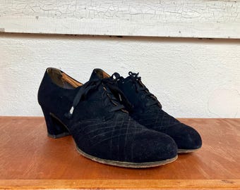 May include: A pair of black vintage shoes with a lace-up design. The shoes have a textured, diamond-patterned vamp and a chunky heel. The shoes are made of a soft, suede-like material.