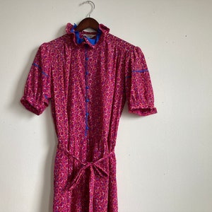 Op de afbeelding: Een roze jurk met bloemenprint, blauwe bies en een strikceintuur. De jurk heeft een gerimpelde kraag en korte mouwen.