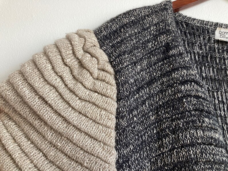May include: A beige and black knitted cardigan with a ribbed texture. The cardigan has a V-neck and a long sleeve. The brand name "Born Bill" is visible on the tag.