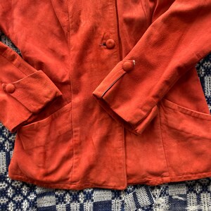 May include: A burnt orange suede jacket with a single button closure and two pockets. The jacket has a slightly fitted silhouette and features a contrasting stitching detail on the sleeves.
