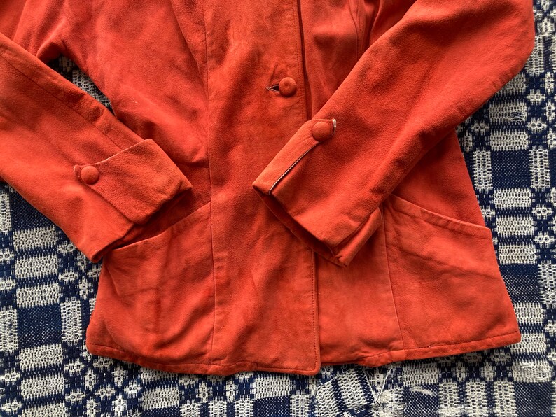 May include: A burnt orange suede jacket with a single button closure and two pockets. The jacket has a slightly fitted silhouette and features a contrasting stitching detail on the sleeves.