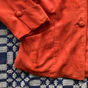 May include: A rust-colored suede jacket with a button closure and a pocket on the left side.