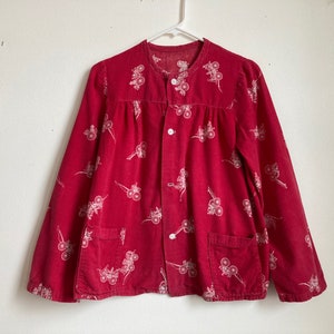 Könnte beinhalten: Ein rotes Cordhemd mit Knöpfen und einem weißen Blumenmuster. Das Hemd hat lange Ärmel und zwei Taschen.
