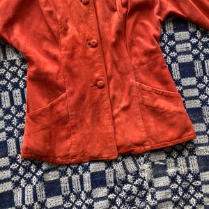 May include: A rust-colored suede jacket with a button-up front and two pockets. The jacket is on a blue and white patterned surface.