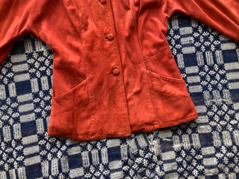 May include: A rust-colored suede jacket with a button-up front and two pockets. The jacket is on a blue and white patterned surface.