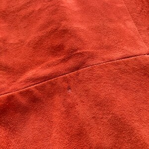May include: Close-up of a rust-colored suede fabric with a small hole in the center. The fabric has a smooth, velvety texture.