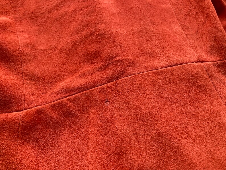 May include: Close-up of a rust-colored suede fabric with a small hole in the center. The fabric has a smooth, velvety texture.